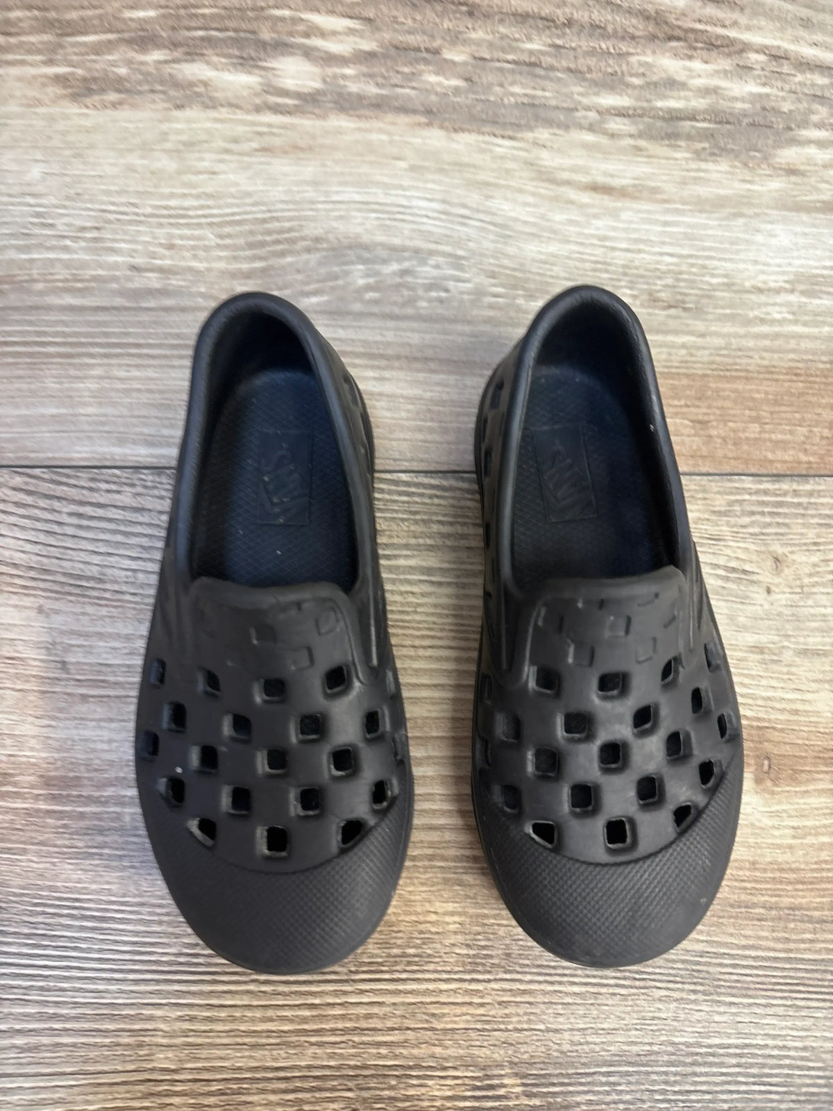 Top-down view of Vans Slip-On TRK toddler black sandals showcasing square ventilation holes and textured toe on wood floor