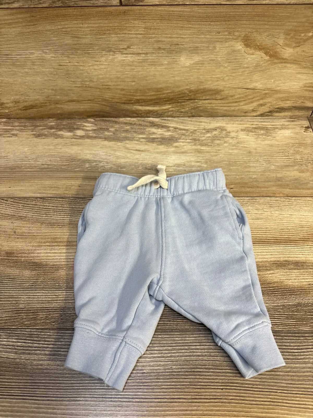 Flat lay of light blue baby joggers (3 months) showcasing elastic drawstring waist, side pockets, and cuffed ankles on wood background