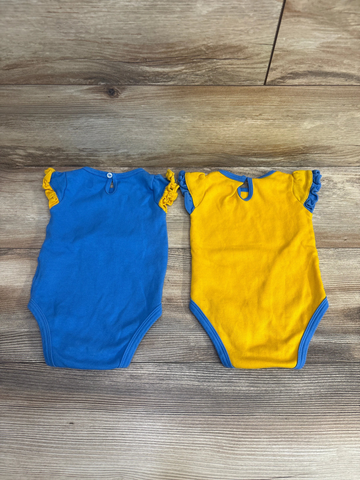 Two colorful baby bodysuits with ruffled sleeves, one blue with yellow accents, one yellow with blue accents, on wooden floor