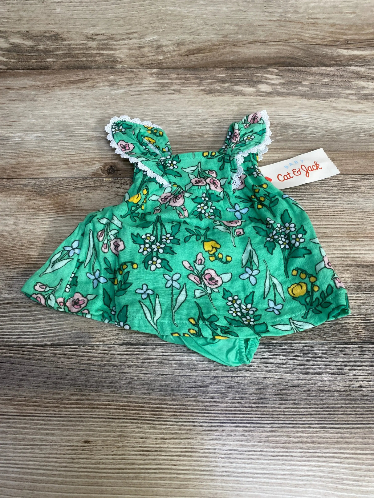 Flat lay of newborn green floral sleeveless dress with matching bloomers, lace-trimmed ruffle straps on wood background