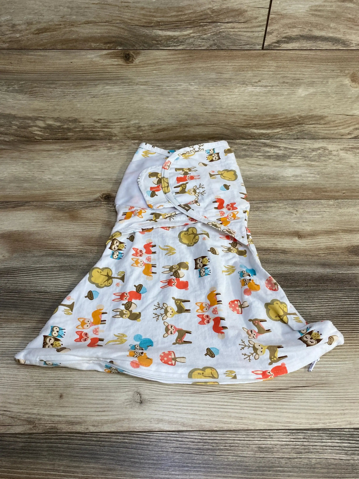 Flat lay of white zippy swaddle (0–3M) showing woodland animal print, front zipper and wrap wings on wood background