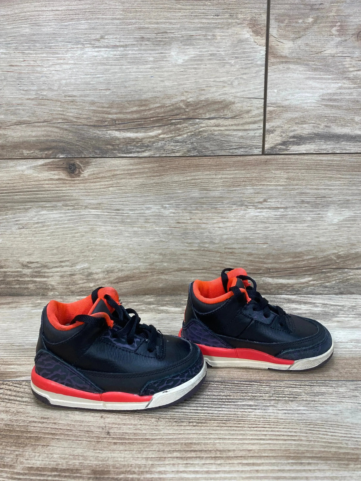 Front-side view of toddler Air Jordan 3 Retro TD sneakers in black with crimson accents and orange-red lining on wood floor