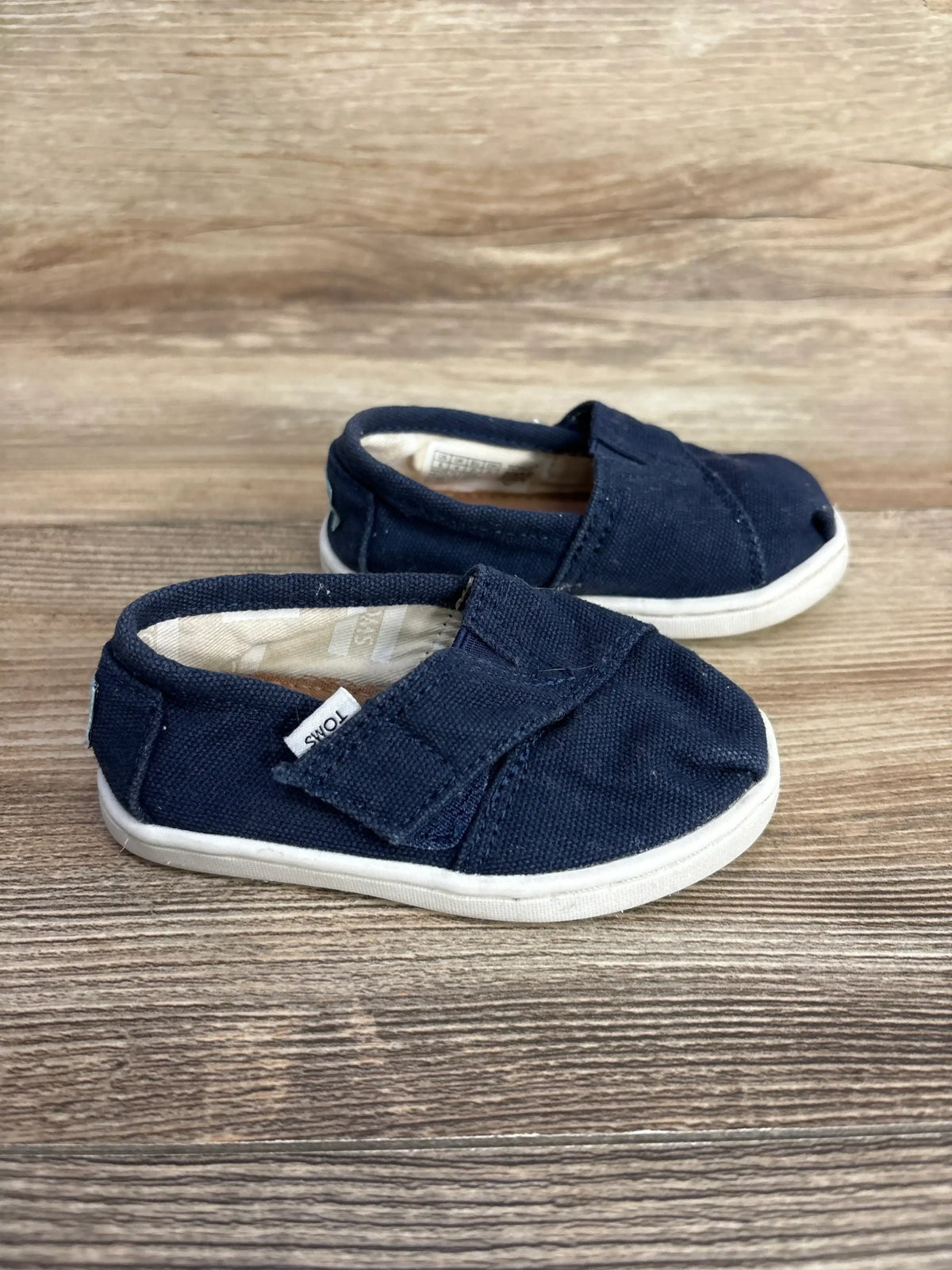 Angled side view of Toms Tiny Alpargata toddler shoes in navy showing canvas upper, Velcro strap, and white soles on wood
