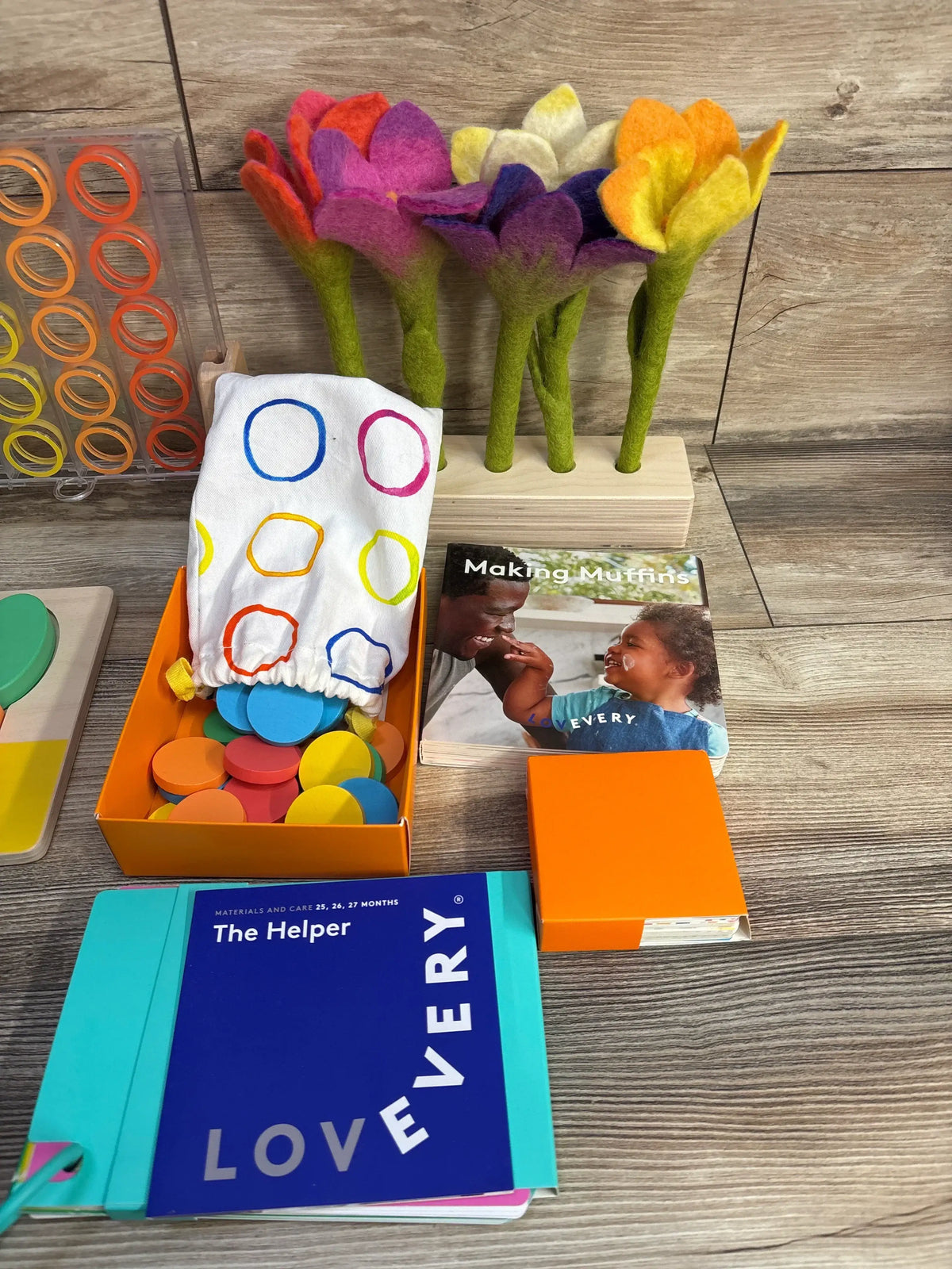 Overhead view of Lovevery The Helper kit with blue guidebook, orange box of multicolored discs, and felt flowers on wood