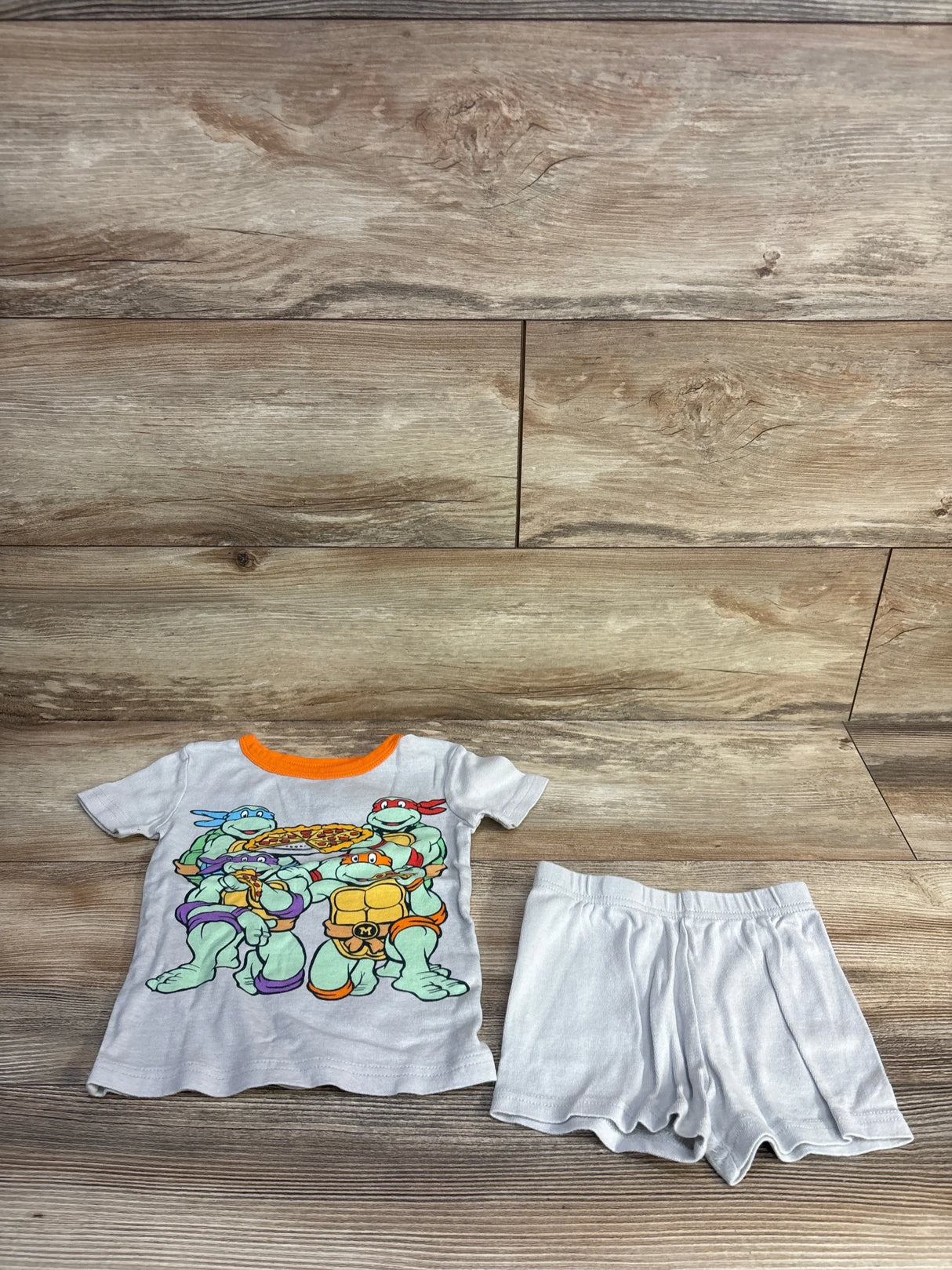 Flat lay of 2-piece Teenage Mutant Ninja Turtles pajama set size 4T with graphic top and white shorts on wood background