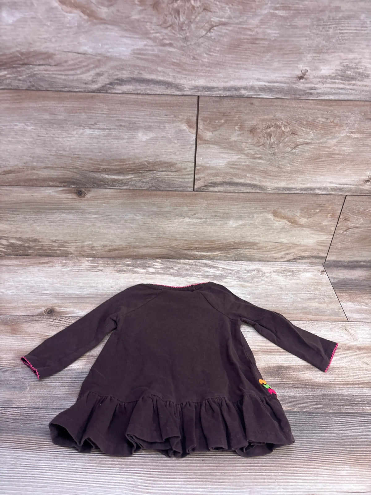 Back view of Bonnie Baby Turkey brown peplum top showcasing ruffled hem and pink trim on a wood-look surface