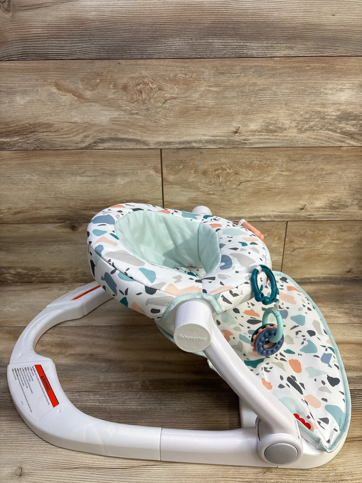 Angled view of Fisher Price Sit-Me-Up Floor Seat in Pacific Pebble showcasing patterned seat, two toys, and wide base on wood