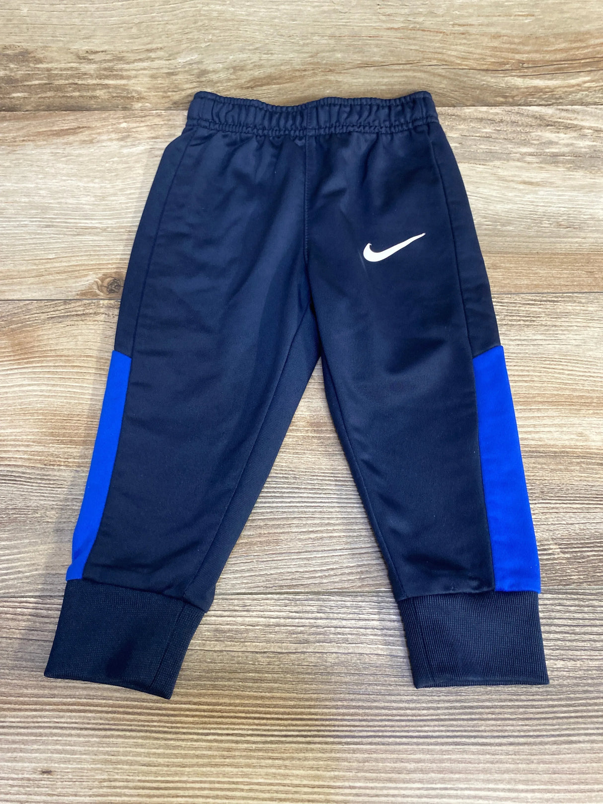 Front view of navy Nike toddler joggers (12 months) showcasing white swoosh, blue side panels, elastic waistband and ribbed ankle cuffs on wooden floor