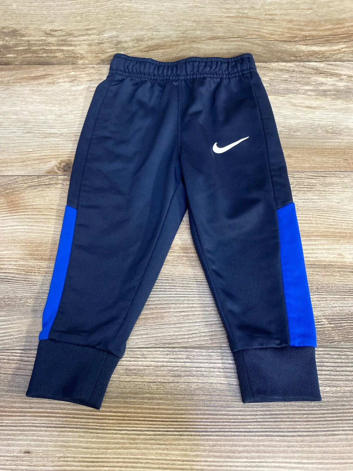 Front view of navy Nike toddler joggers (12 months) showcasing white swoosh, blue side panels, elastic waistband and ribbed ankle cuffs on wooden floor