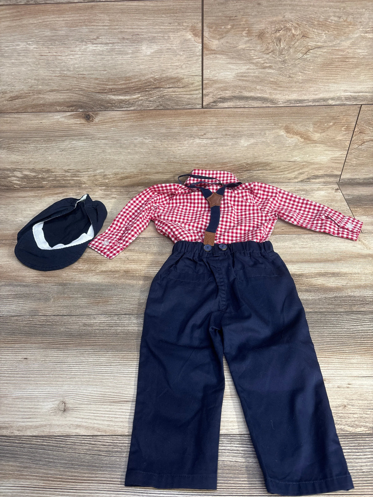 Children's outfit with red checkered shirt, navy blue pants with suspenders, and navy cap on wood floor