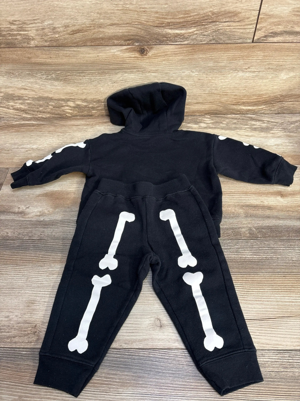 Top view of Child Of Mine 2pc skeleton hoodie and joggers in black, size 12m, with white bone graphics on a wood floor