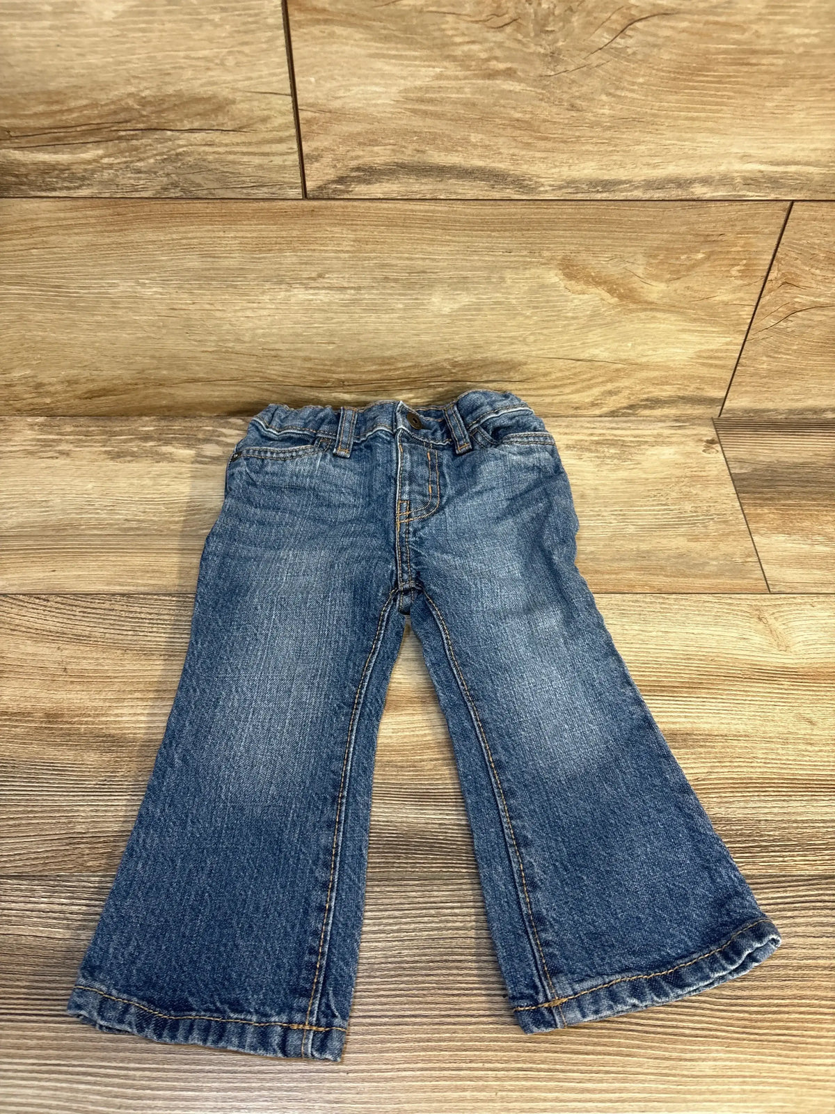 Top-down front view of OshKosh light blue boot-cut toddler jeans, 18 months, elastic waistband and faded wash on wood floor