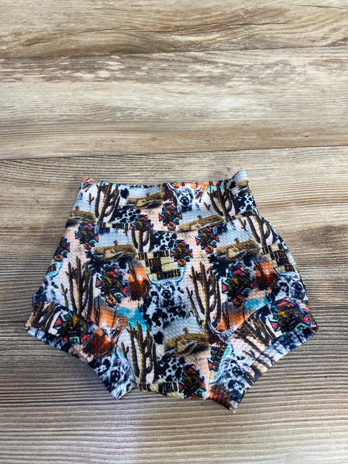 Colorful baby shorts with western print including cacti, cowboy hats, and dogs on wood background