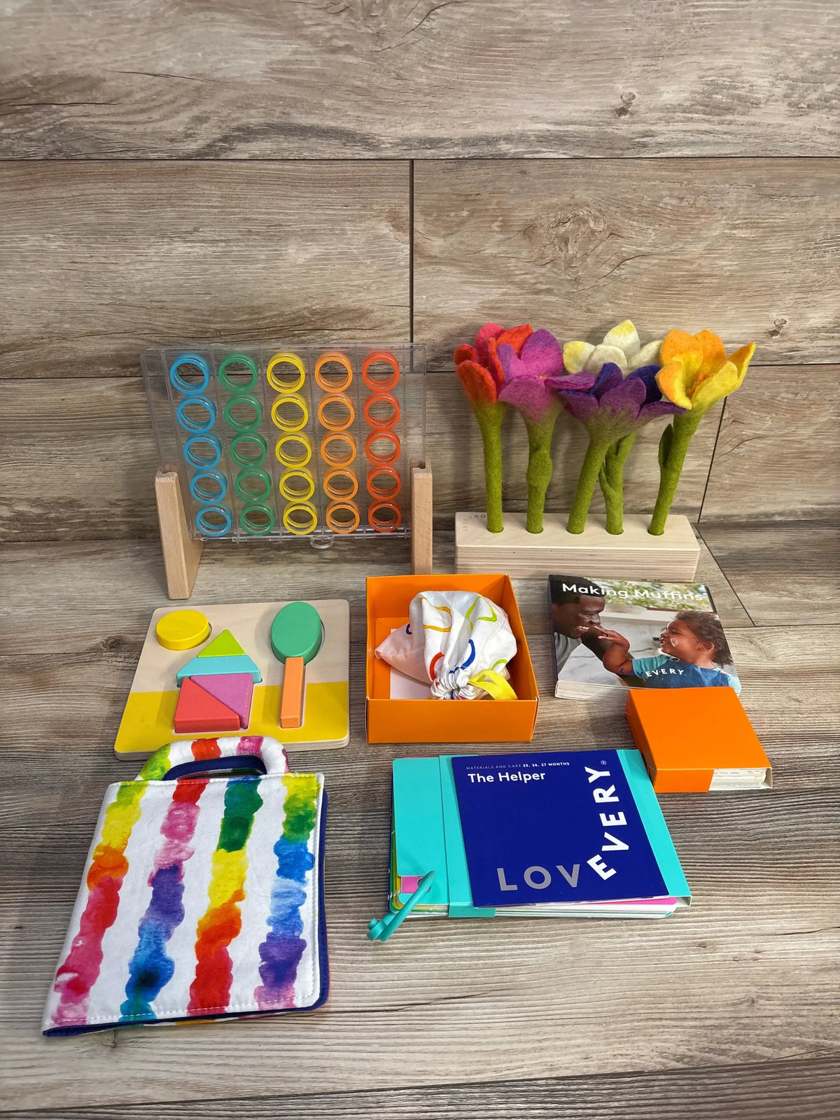 Front view of Lovevery The Helper play kit with activity board, wooden puzzle pieces, felt flowers and cloth book