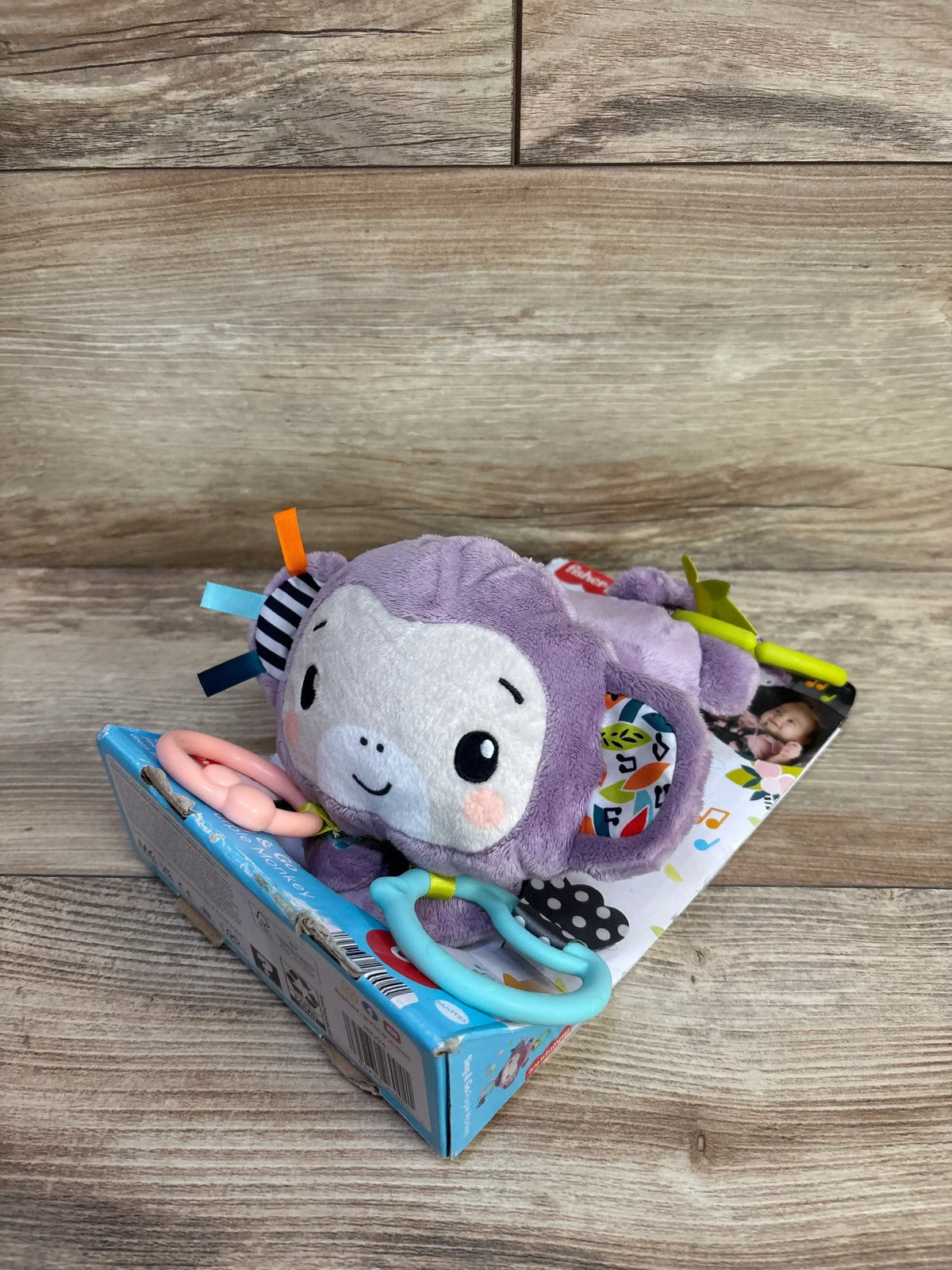 Angled view of Fisher-Price Sing and Go purple monkey stroller toy showcasing plush sensory details, clackers, ribbons, and link on retail box
