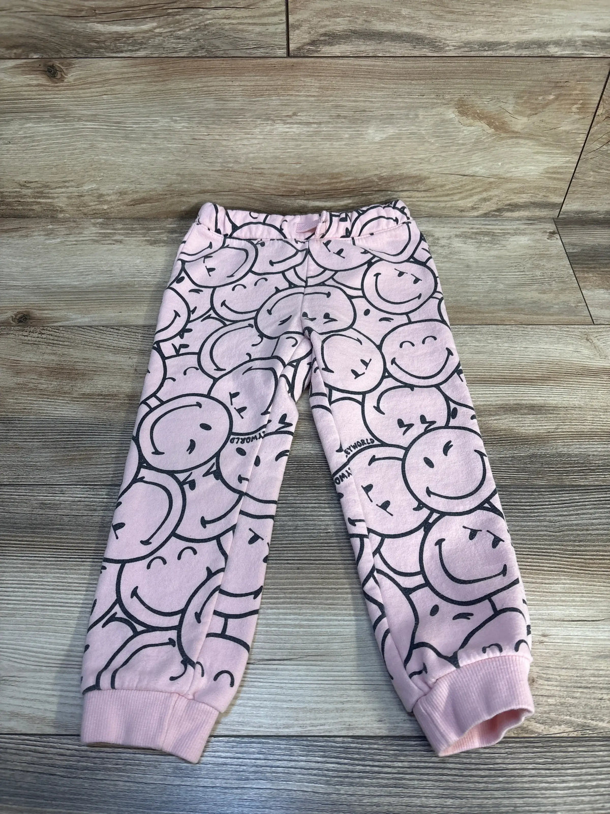 Flat lay of pink 4T toddler joggers showing black smiley-face print, drawstring waist, and ribbed ankle cuffs on wood floor