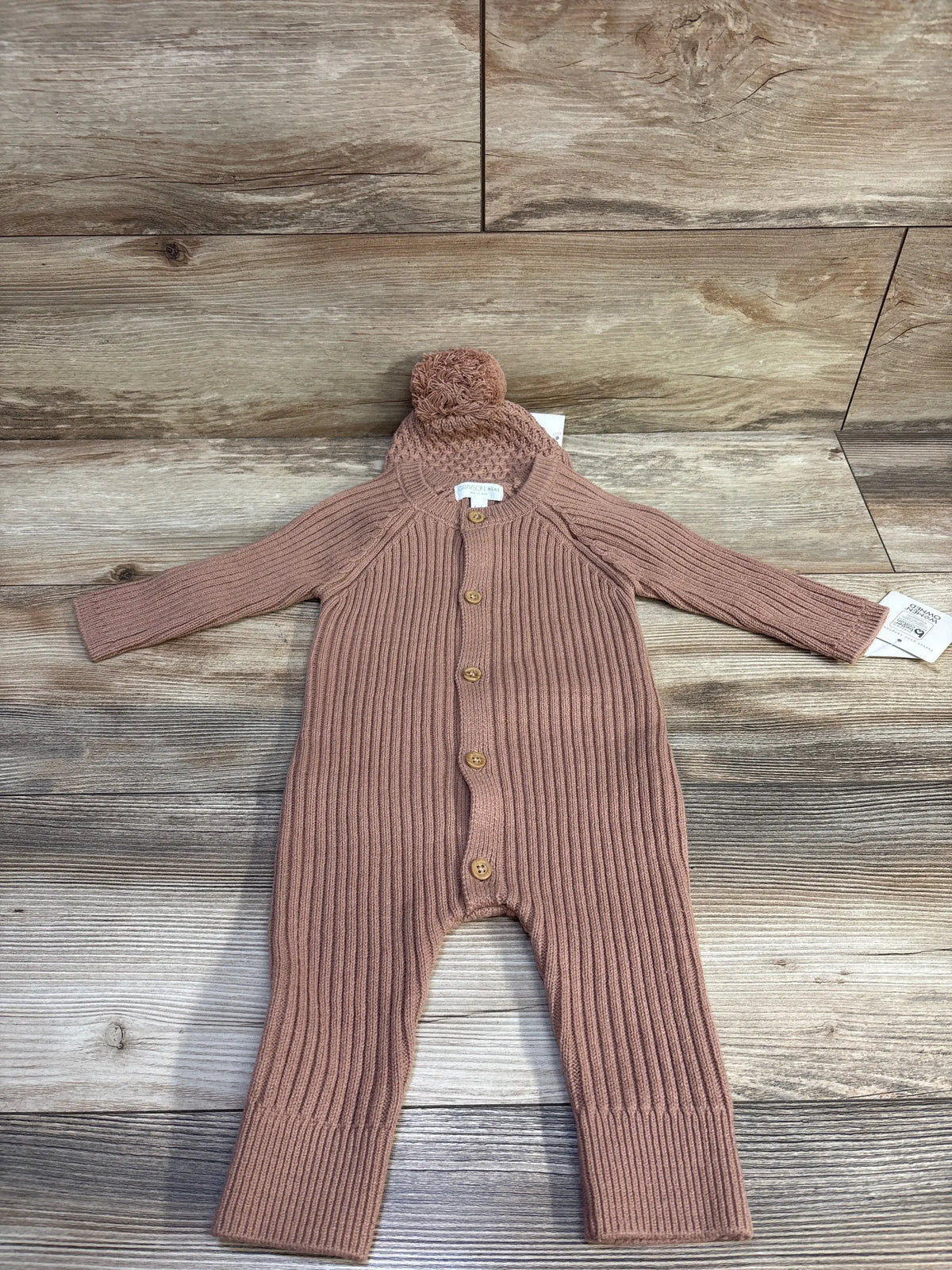 Flat lay of Grayson Mini knit coverall and pom-pom hat set showcasing ribbed texture and wooden buttons on light wood background
