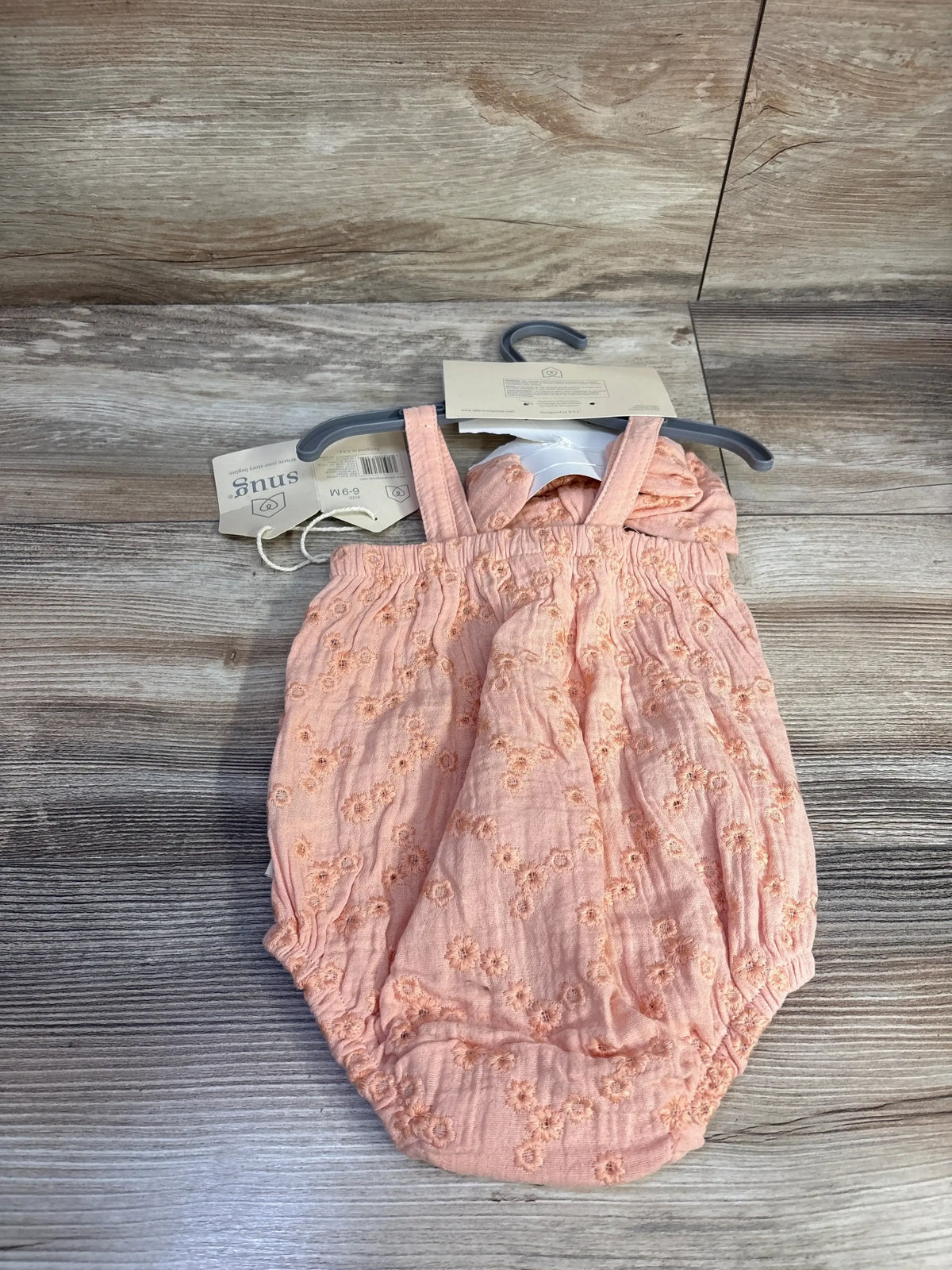 Top view of peach embroidered bubble romper with matching headband on hanger showcasing floral eyelet embroidery and elasticized leg openings on wood background