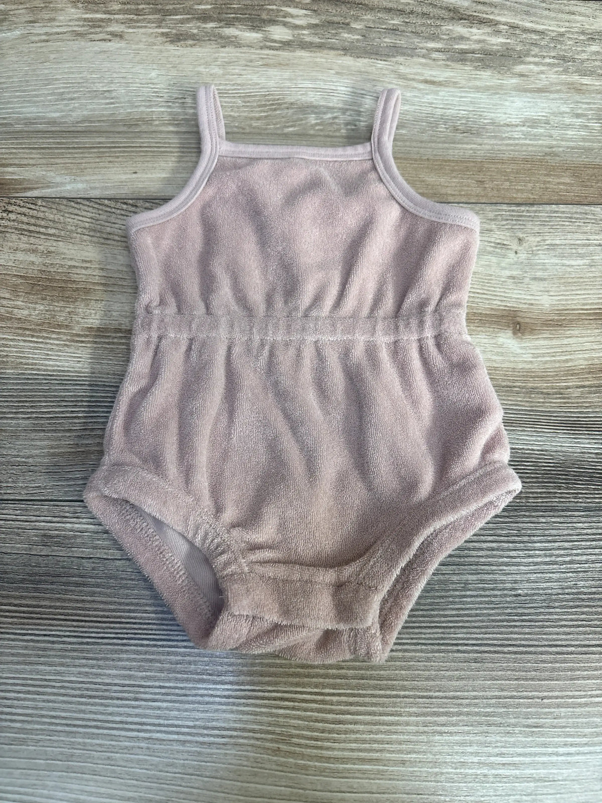 Flat lay of Quincy Mae terry cinch romper in dusty pink showing cinched waist, thin shoulder straps, and terry cloth texture on light wood background