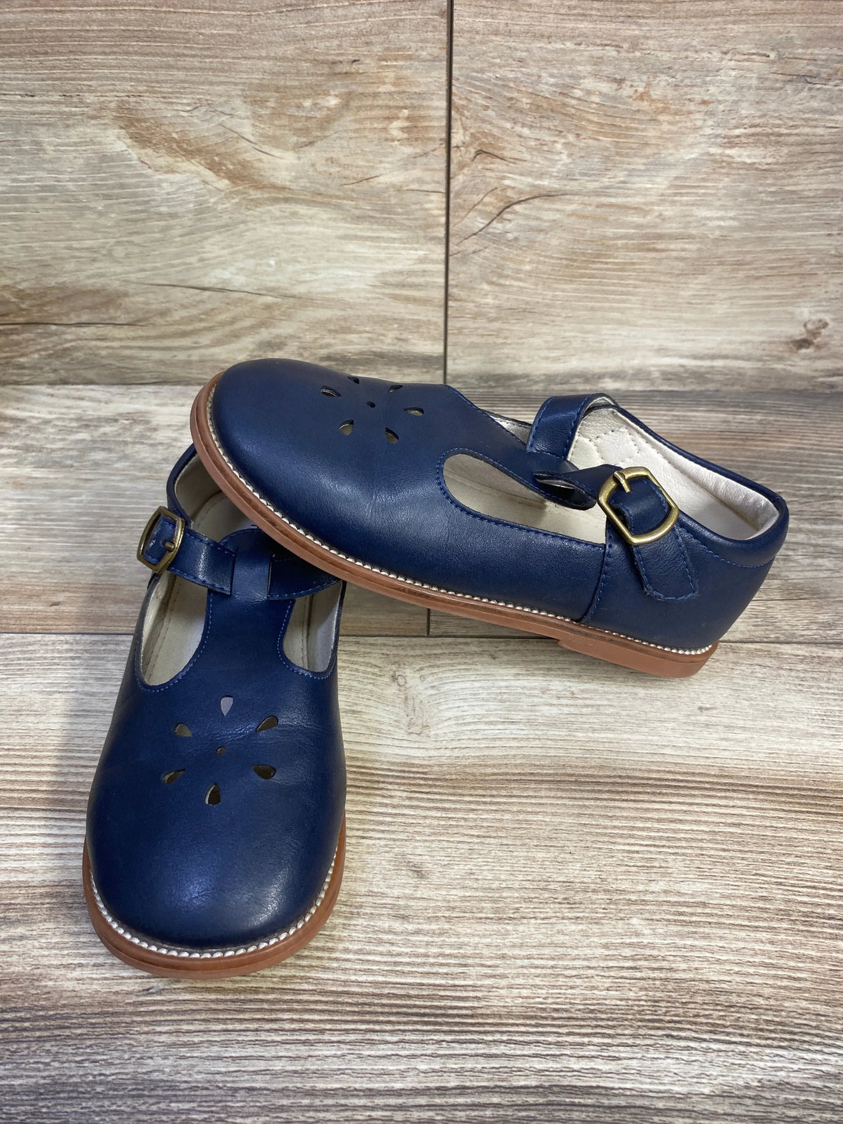 Angled view of Felix & Flora navy T-strap Mary Jane flats showing flower cutouts, gold buckle, and brown sole on wood floor