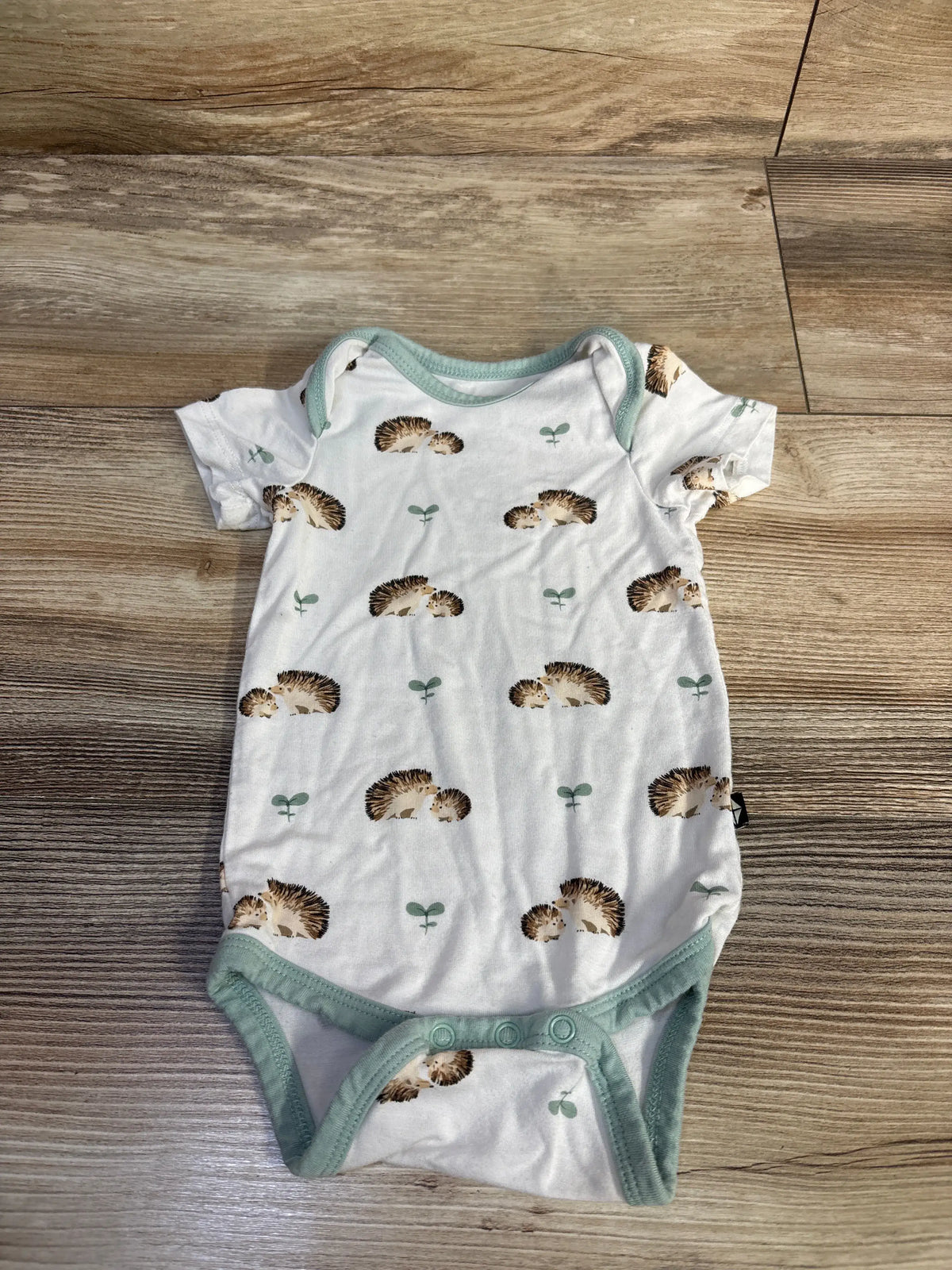 Front view of Kyte Baby bodysuit in Hedgehog White, size 0-3 months, with hedgehog print, green trim, and snap closures on wood