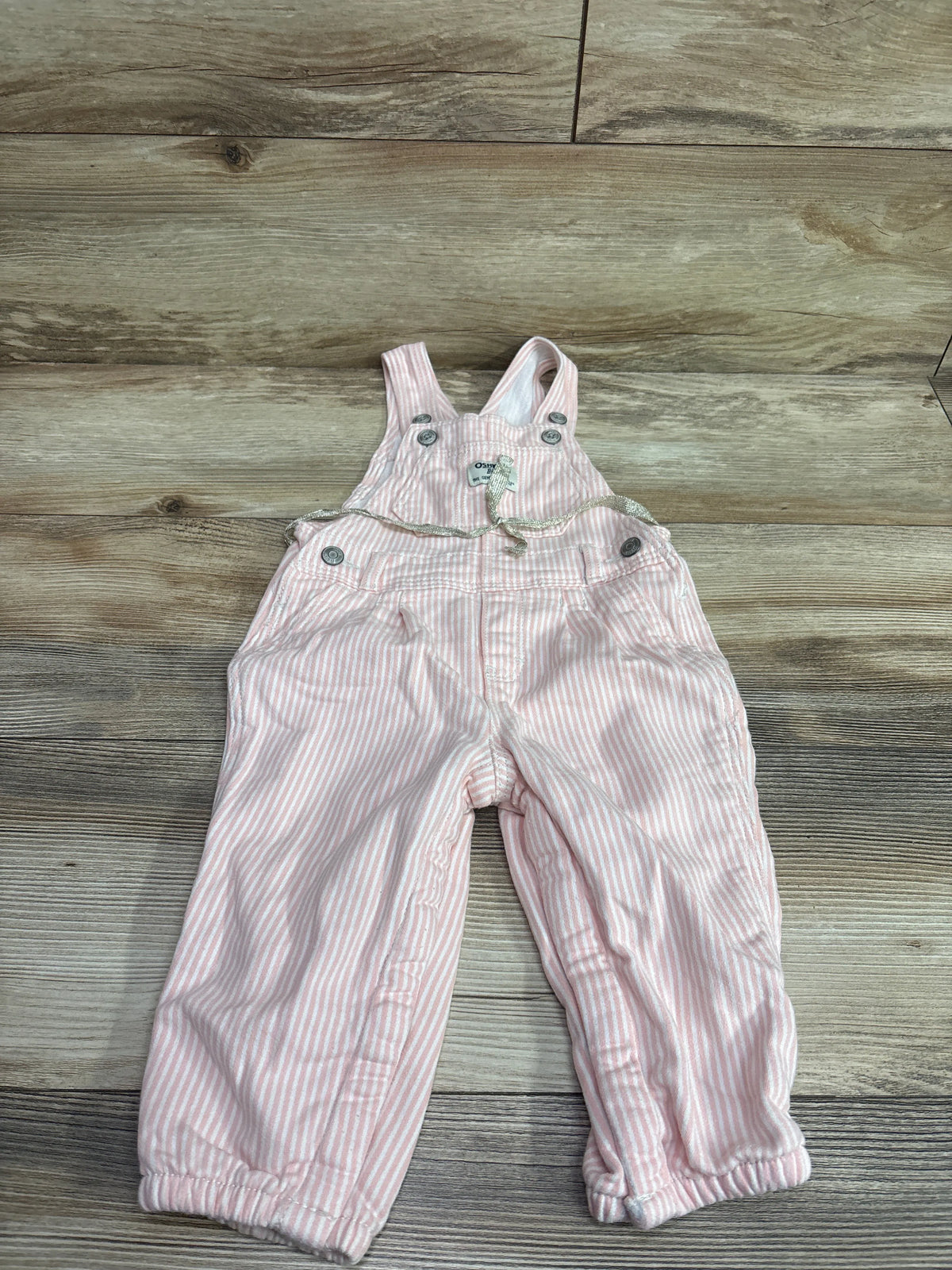 Front view of Oshkosh pink and white striped overalls in size 18 months with bib straps, buttons, pockets, and elastic cuffs on a wood floor