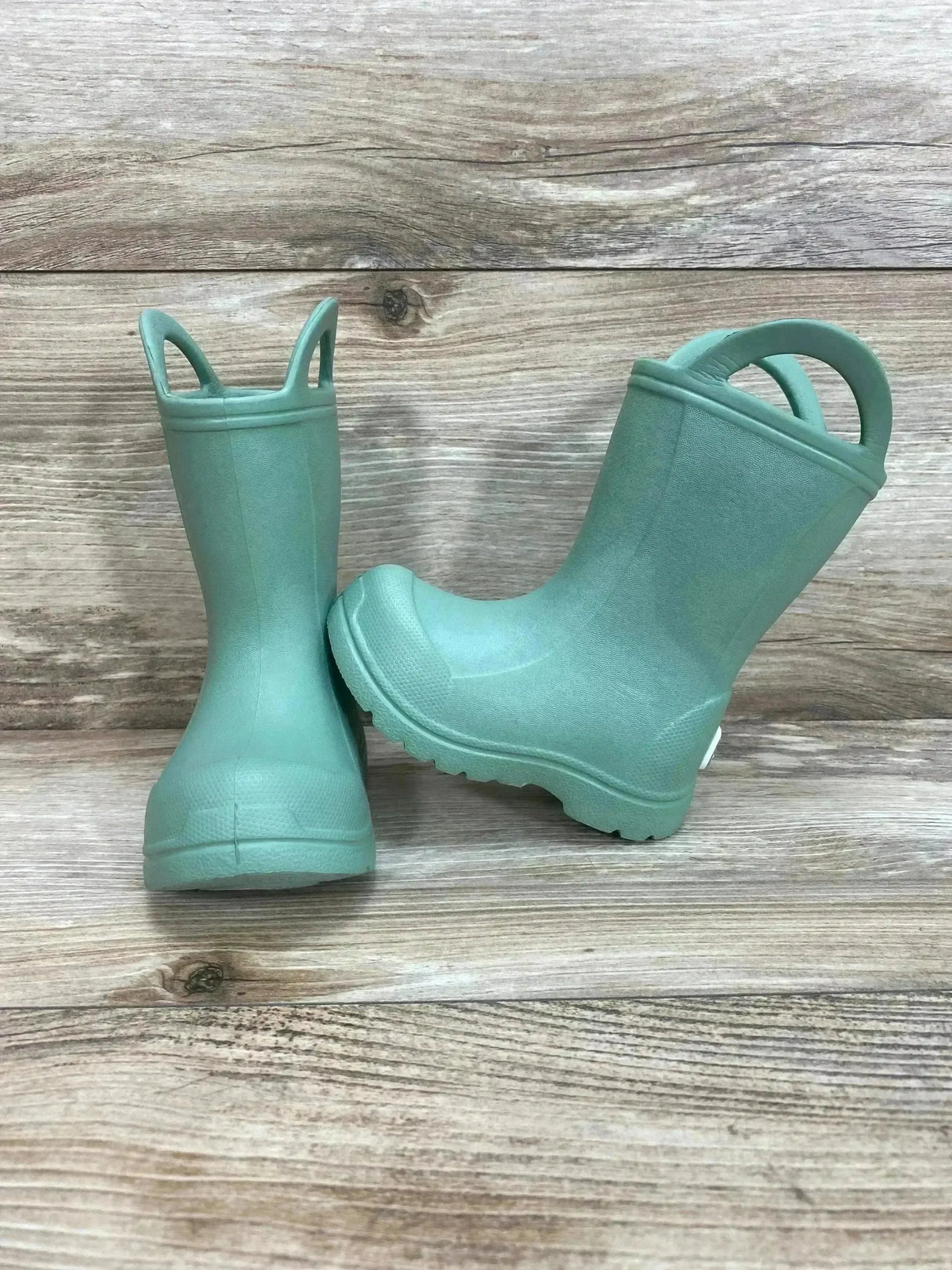 Front and side view of green BEARPAW toddler EVA rain boots with pull on handles and lugged tread on light wood background