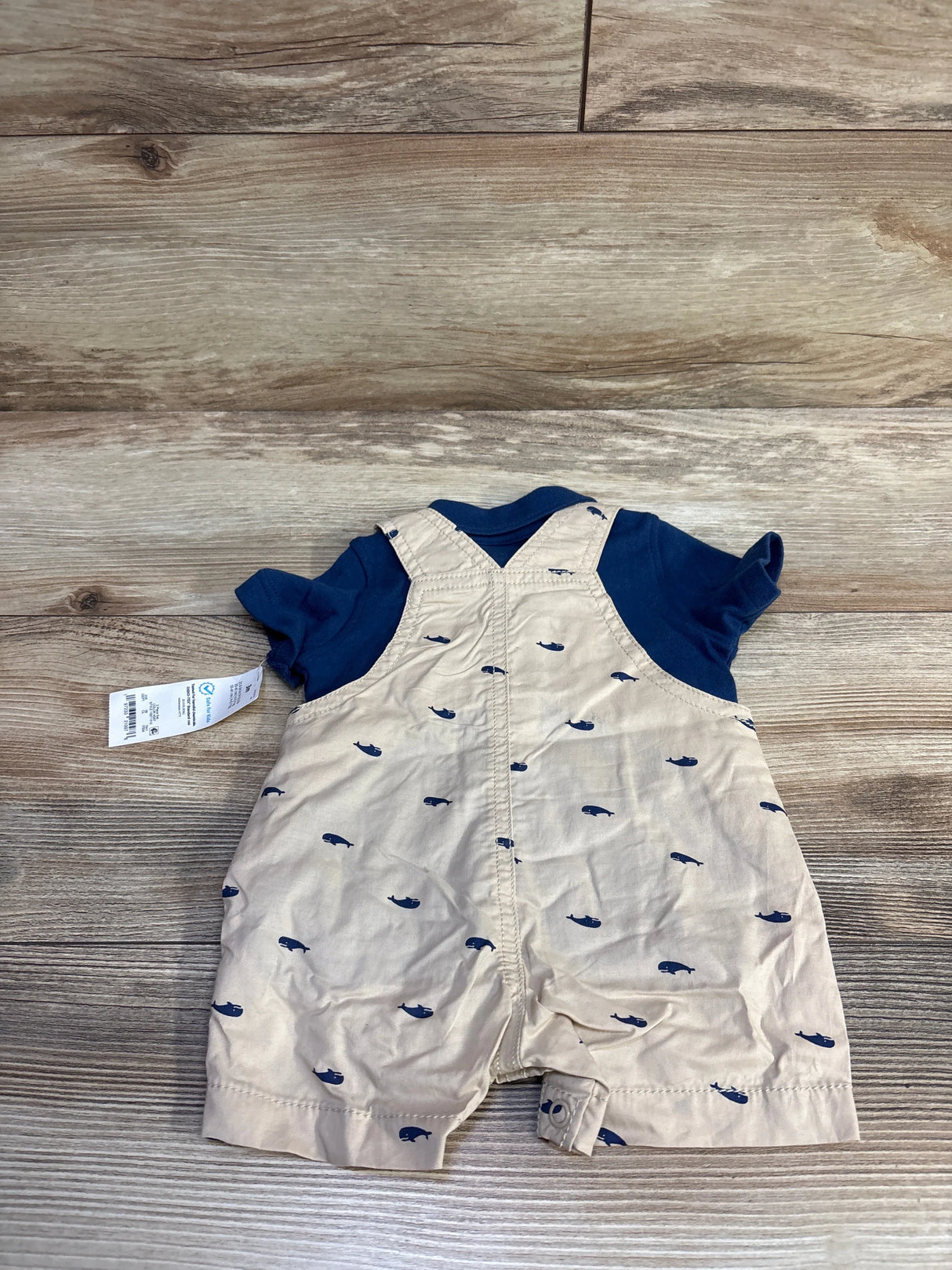 Flat-lay view of baby polo and shortalls set showing navy polo and beige shortalls with navy whale print on wood background
