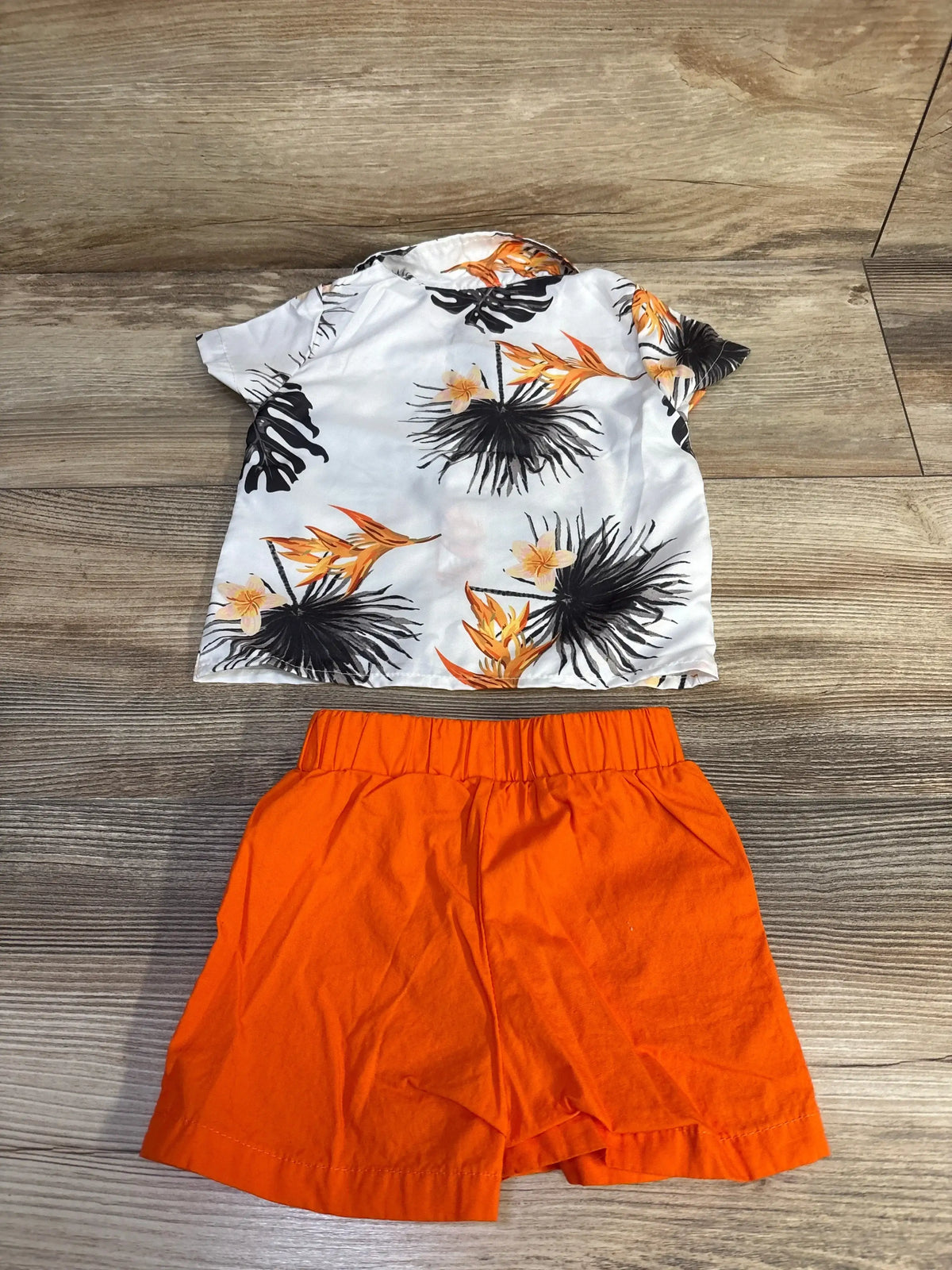 Flat lay of infant 2 piece: white button up shirt with black and orange tropical print and orange shorts on wood floor