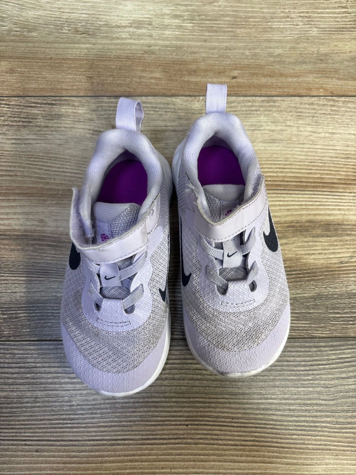 Top view of Nike Revolution 6 TD sneakers showing violet frost upper, black Swoosh, Velcro strap, purple insoles on wood