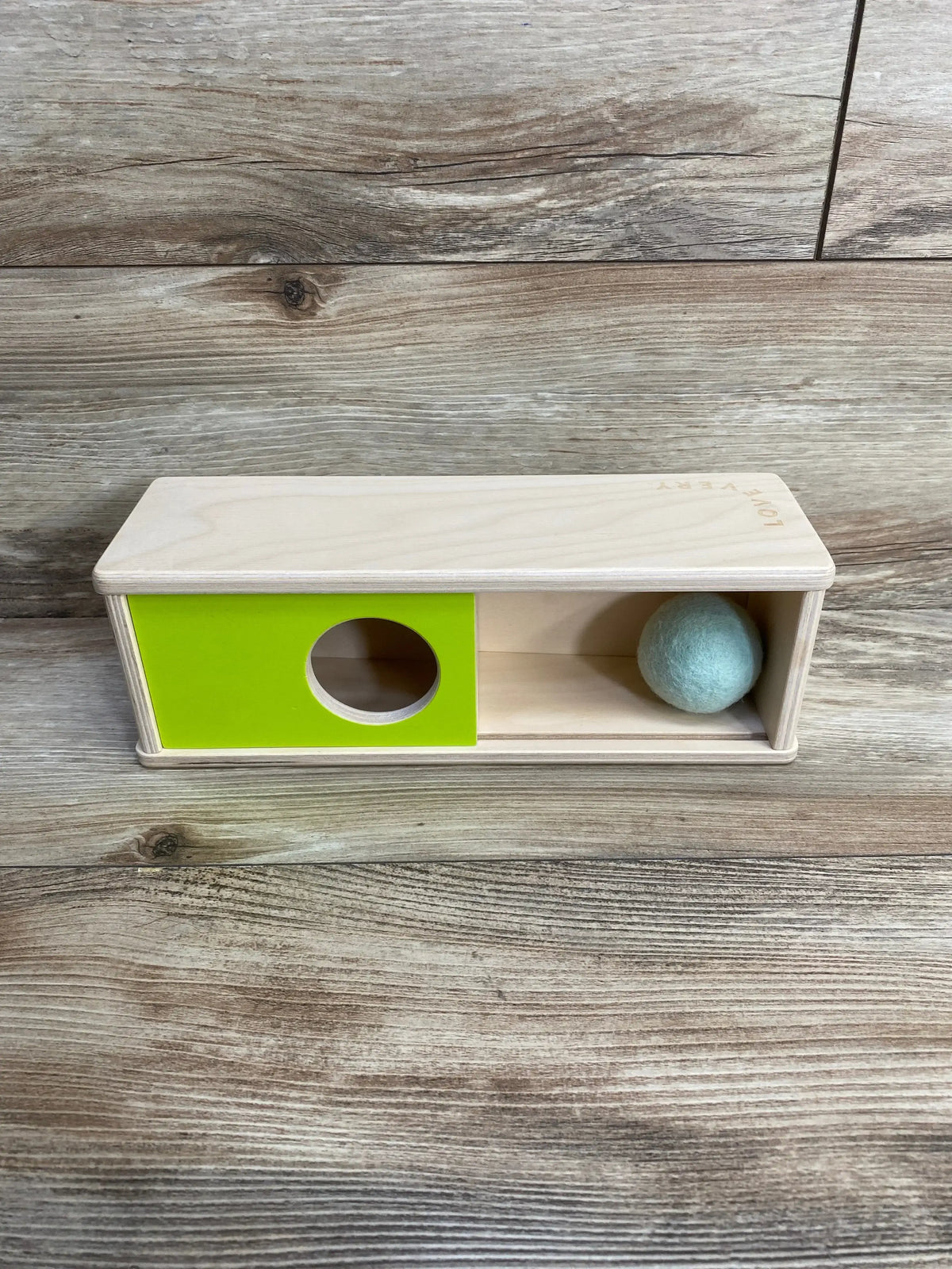 Front view of Lovevery Sliding Top Box showcasing green sliding panel with circular hole and light blue felt ball on wooden surface