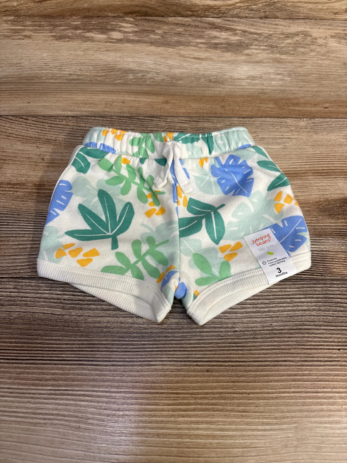 Top view of white Jumping Beans baby shorts with green and blue leaf print, drawstring waist and 3m tag on wood background