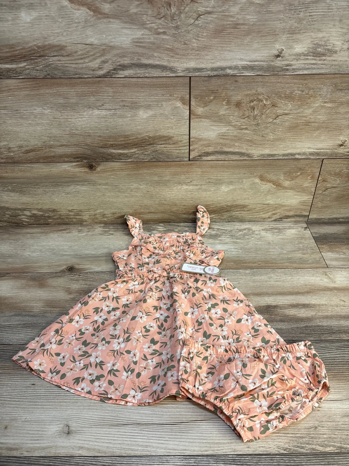 Flat lay of Little Lass peach floral toddler dress and matching bloomers showing ruffle shoulder straps and smocked bodice on wood background