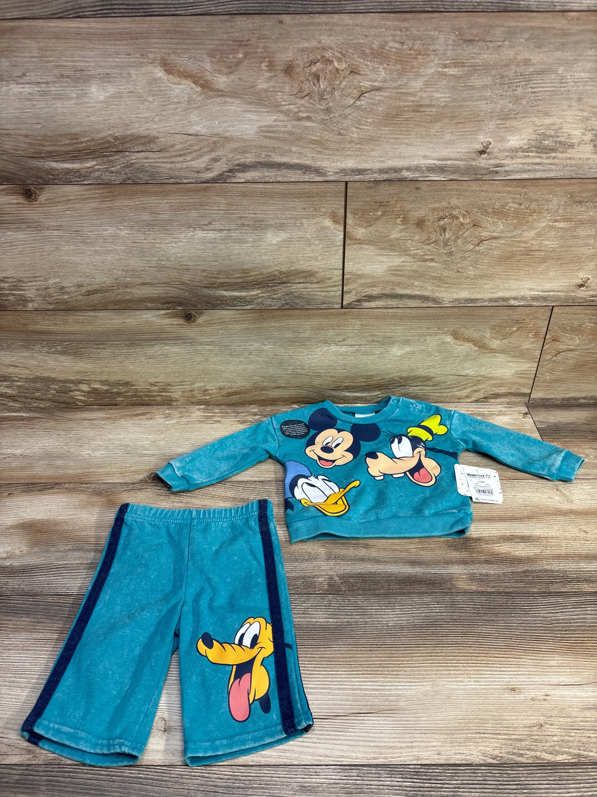 Top view of teal Disney baby sweatshirt and shorts with Mickey, Donald and Goofy on the top and Pluto on the shorts on wood