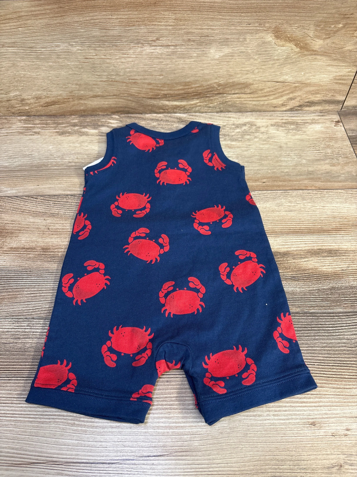 Flat lay of navy Henley shortie romper (6M) showing red crab print, sleeveless Henley front and snap crotch on wood background