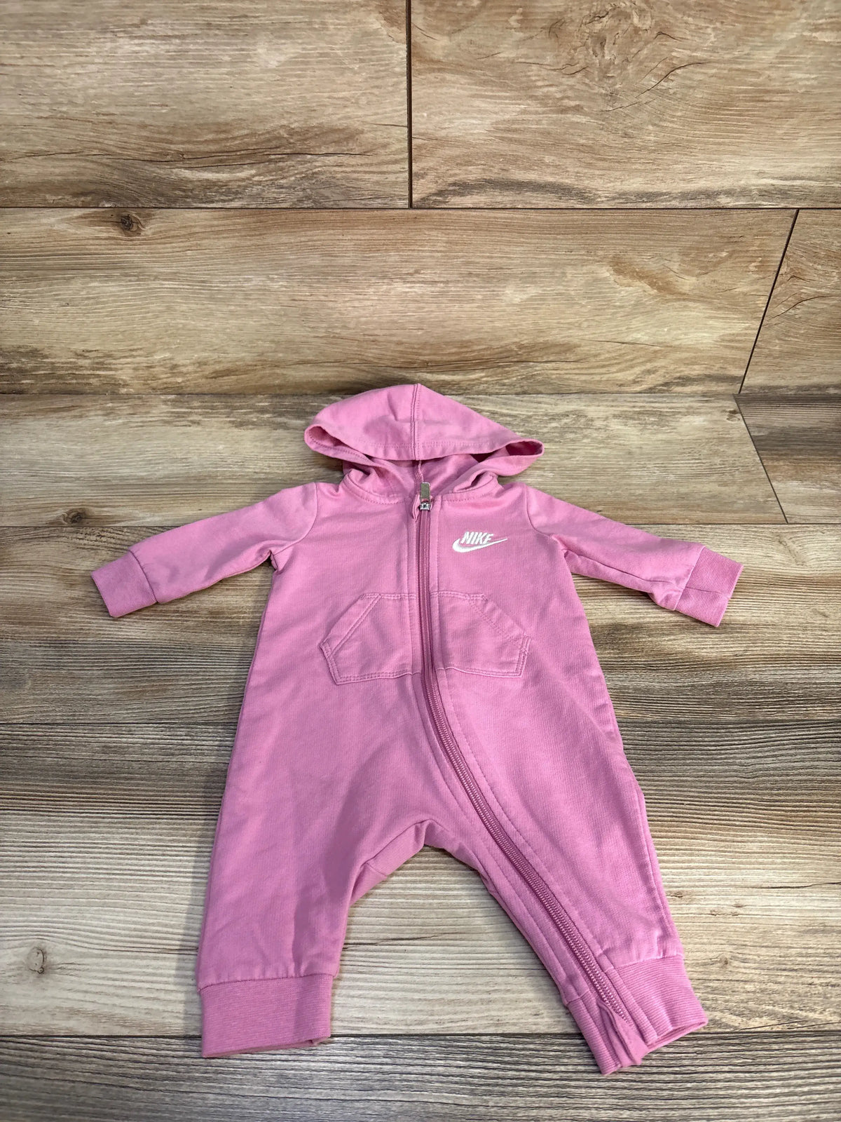 Front view of a pink Nike hooded coverall for 3 months showcasing hood, full-length zipper, and two pockets on a wood floor