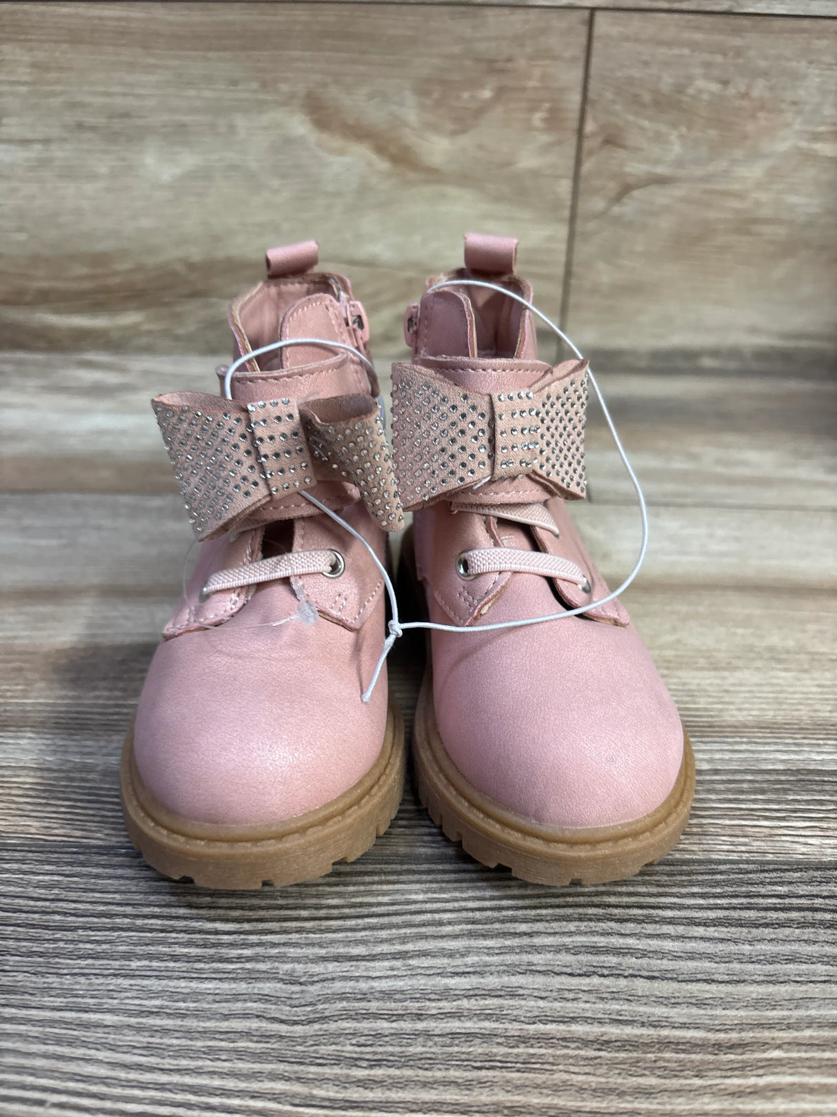 Front view of Daria bow ankle boots in pink showcasing rhinestone studded bows and tan lug soles on wood floor