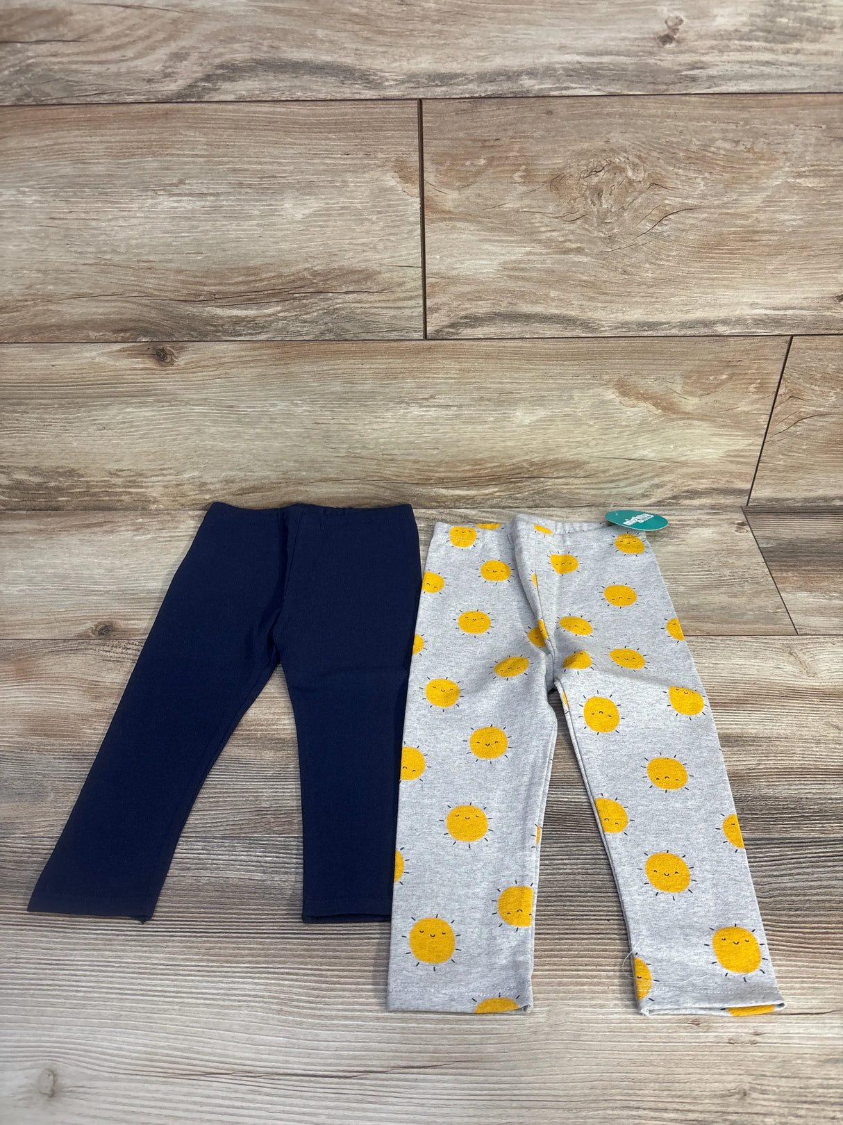 Flat lay of 2-pack toddler leggings, one solid navy and one light grey with yellow smiling sun print, size 18 months on wood floor background
