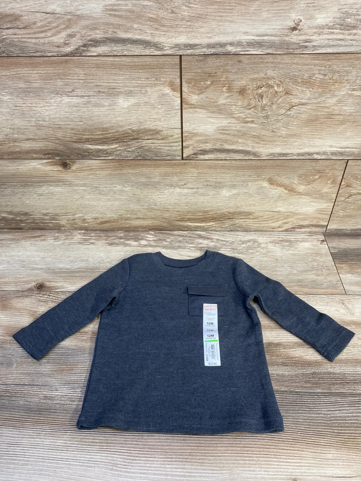 Flat lay of Jumping Beans long sleeve baby shirt showcasing ribbed knit texture, chest pocket, and visible size tag on light wood background