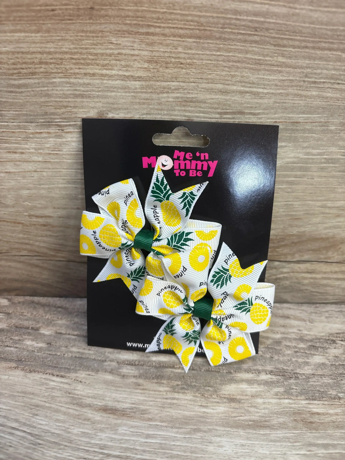 Front view of NEW fanned out hair bows 2 pack with yellow pineapple print ribbon on Me n Mommy To Be card on wood plank floor