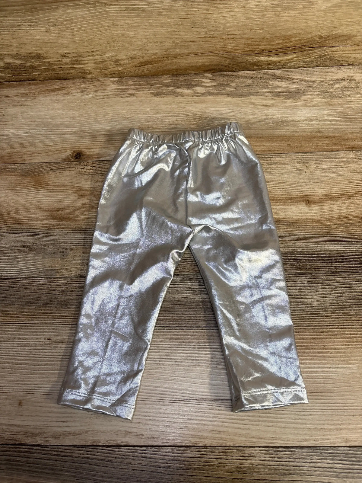 Top-down view of Baby Gap metallic pull-on pants size 12-18 months showcasing silver shiny fabric and elastic waistband on wooden floor