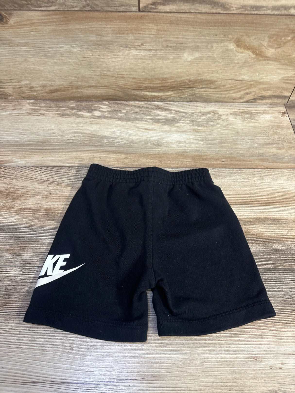 Flat lay of black Nike cotton toddler shorts (24 months) showing elastic waistband and white swoosh logo on wood background