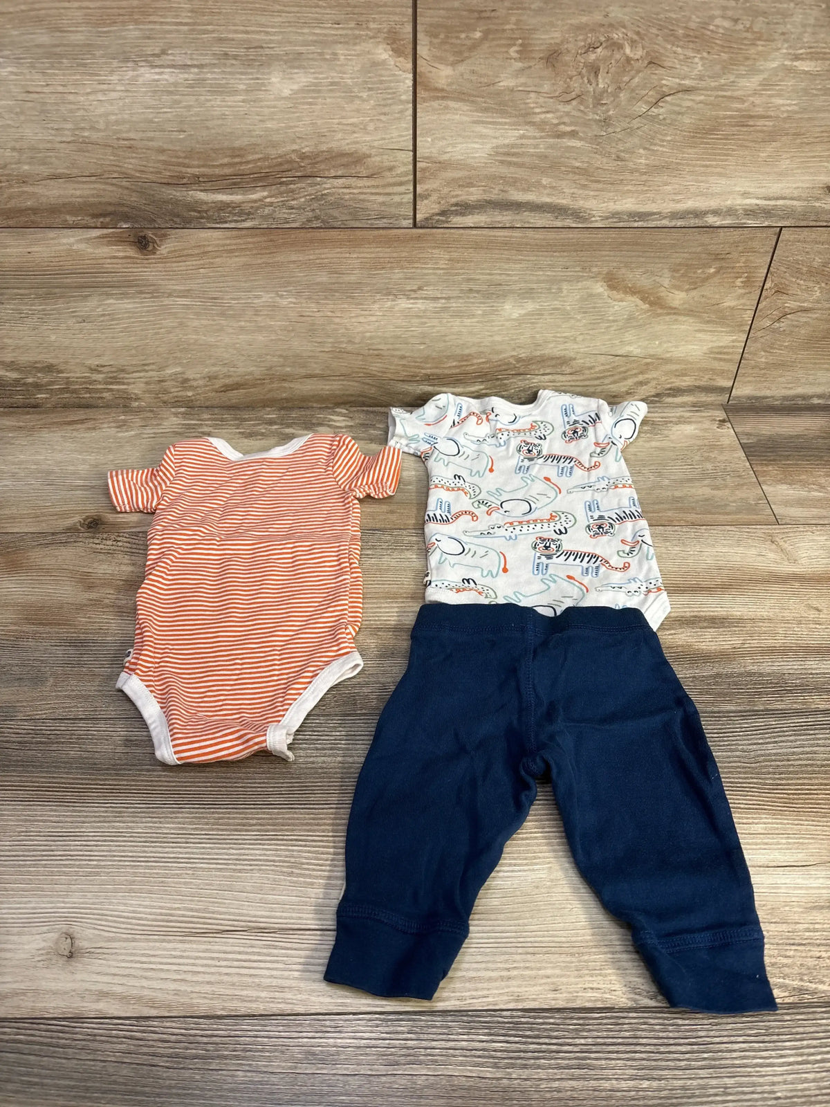 Flat lay of Huggies 3-piece bodysuit set: orange striped bodysuit, white animal-print bodysuit, and navy pants on wood floor