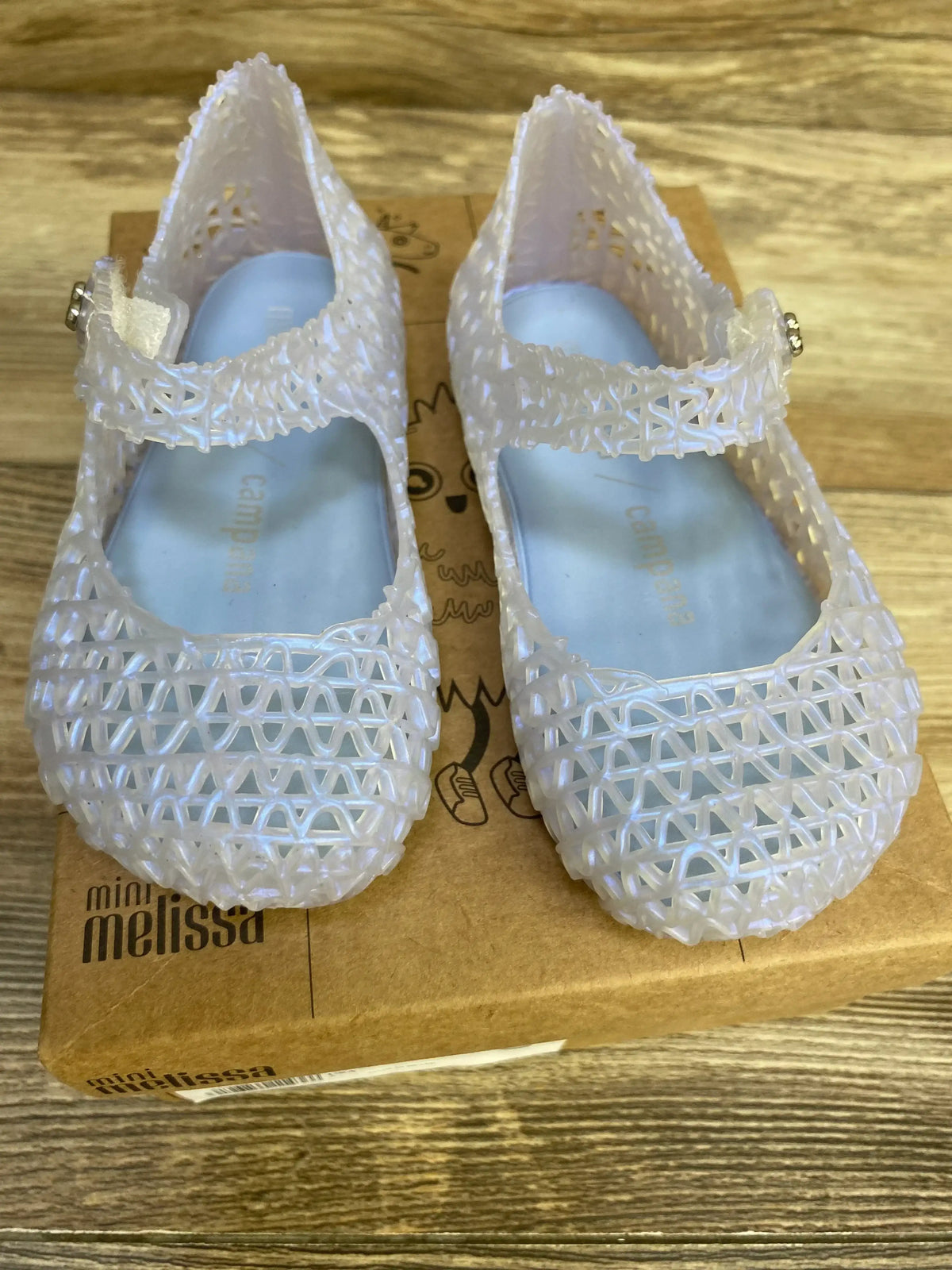 Top-down view of Mini Melissa Campana Papel jelly flats in pearly blue showcasing lattice jelly upper and Mary Jane strap on cardboard shoebox