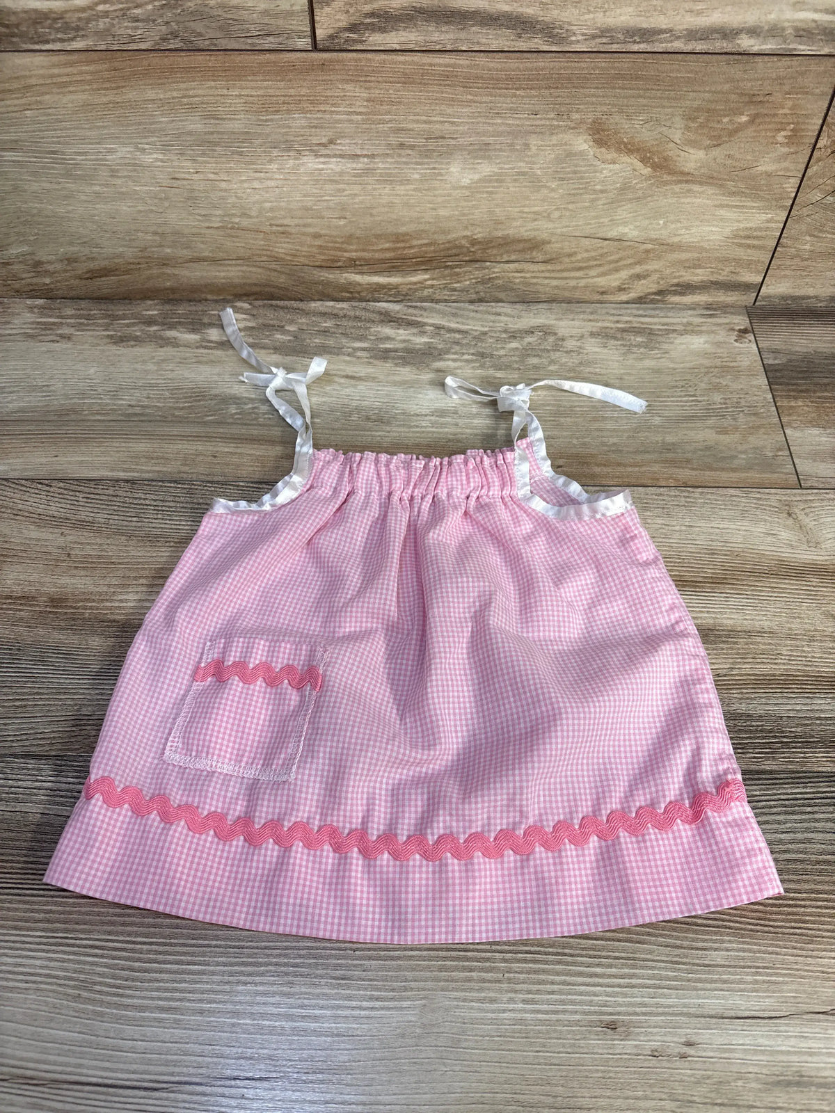 Front view of pink sleeveless gingham sundress in size 6-9m showcasing tie straps, front pocket, and rickrack trim on wood floor