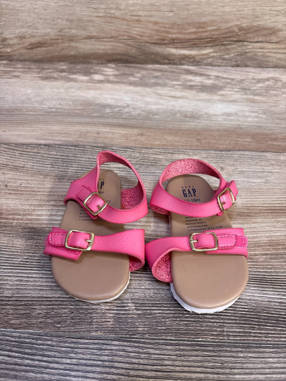 Top-down view of Baby Gap pink toddler buckle sandals showcasing two adjustable gold buckles and tan footbed on wood-grain background