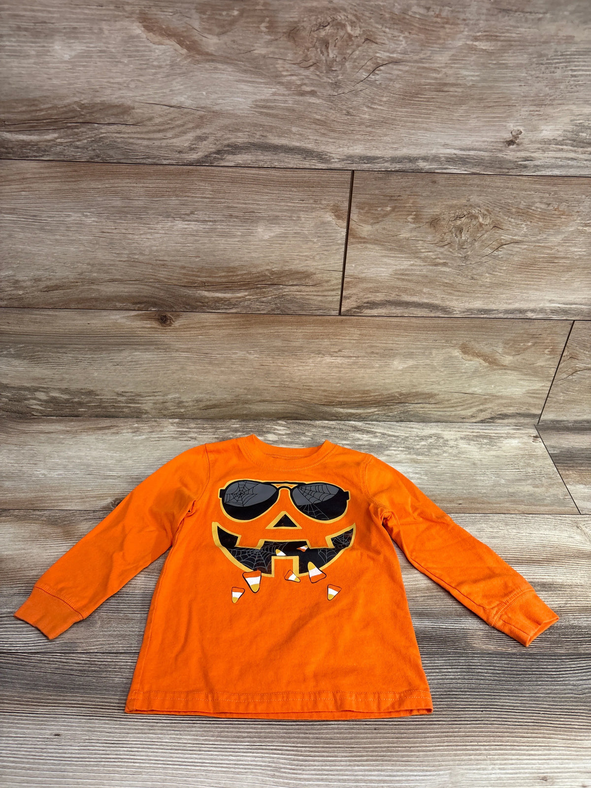 Front view of Carters jack o lantern shirt showcasing orange long sleeves and a sunglasses pumpkin graphic on a wood surface