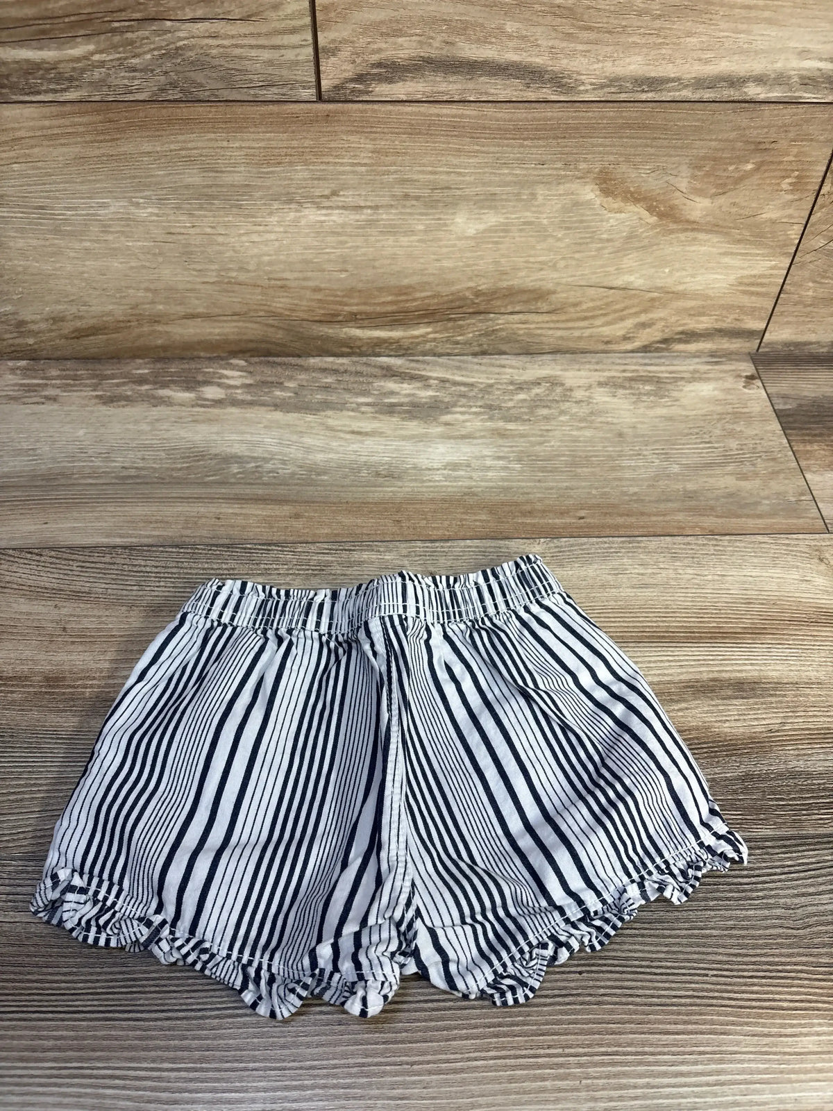 Top-down view of Baby Gap white and navy striped toddler shorts showcasing elastic pull-on waistband and scalloped ruffle hem on wood background