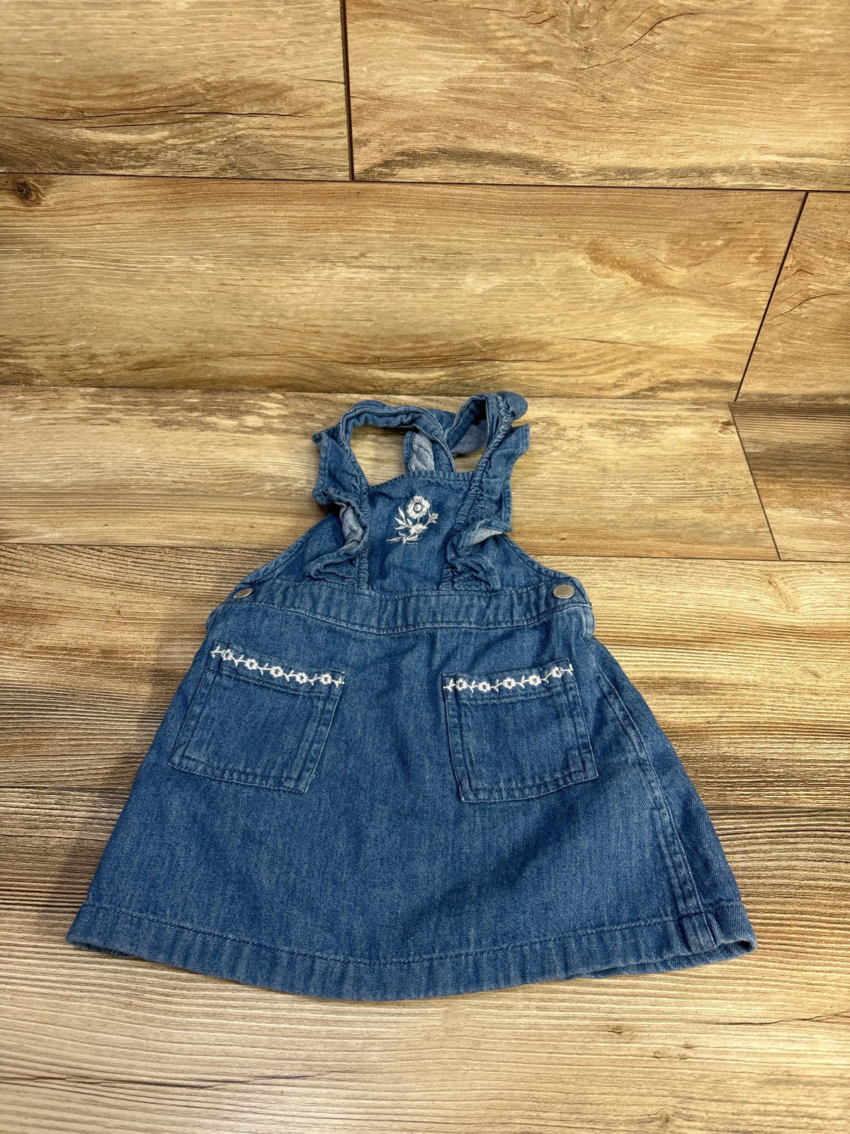 Front view of Old Navy embroidered blue denim skirtall with floral bib embroidery and embroidered pocket trim on a wood floor