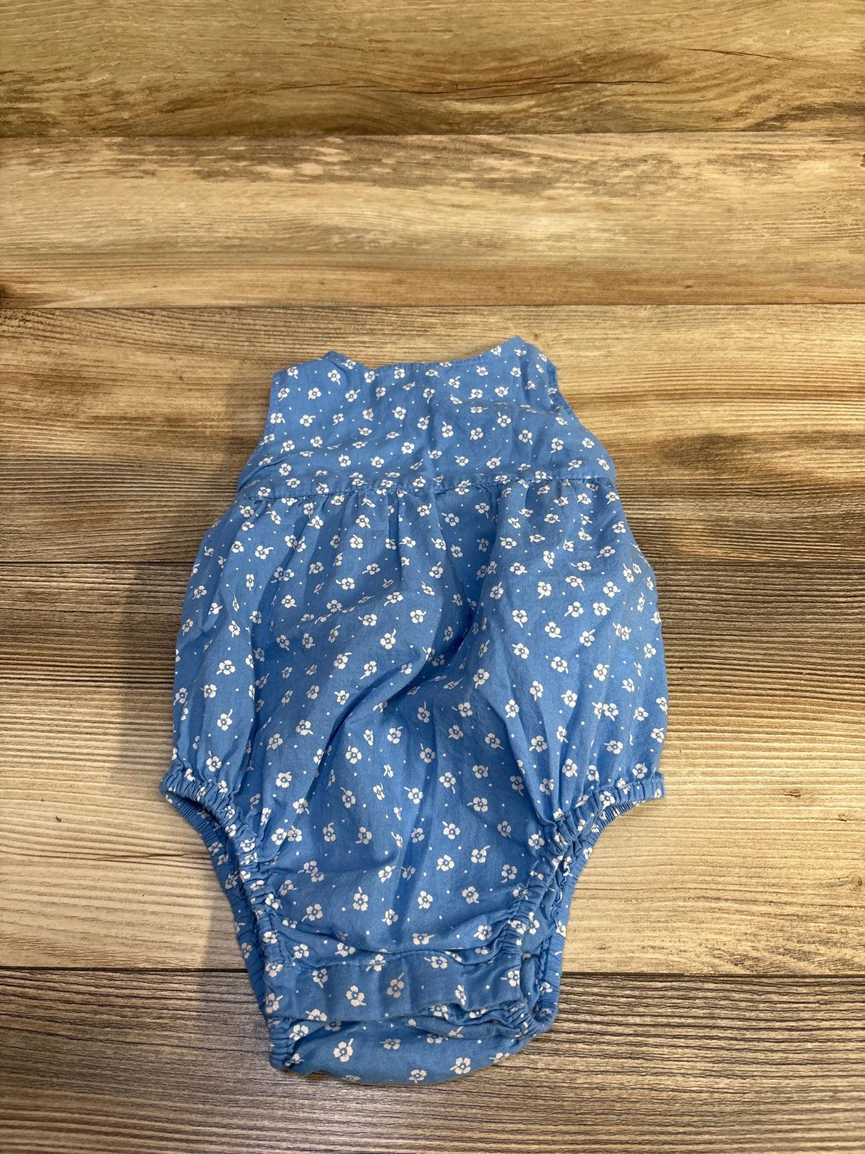 Top view of blue sleeveless bubble romper with white floral print, gathered waist and elastic leg openings on wood background
